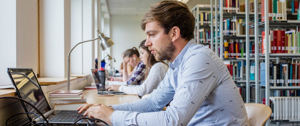 Junger Mann sitzt an seinem Laptop in einer Bibliothek. Hinter ihm, zum Teil nur unscharf zu erkennen, andere Lernende und Bücherregale.