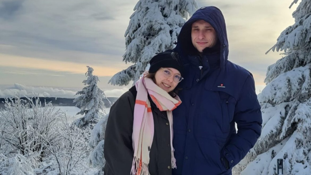 Ein junges Paar steht Arm in Arm in einer verschneiten Winterlandschaft und lächelt in die Kamera.