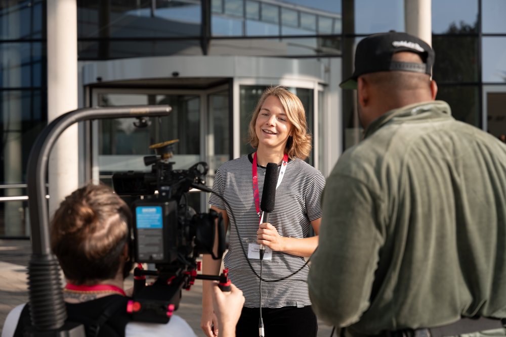 Eine junge Frau steht vor einem modernen Vorlesungsgebäude mit Glasfront, dem Flensburger audimax. Sie spricht in ein Mikrofon, während zwei junge Männer sie mit einer Kamera filmen.