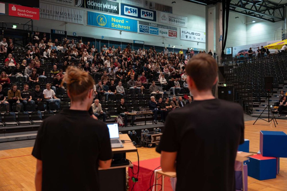 Zwei junge Männer in schwarzen T-Shirts von hinten fotografiert - sie stehen auf einer Bühne mit Blick auf eine gut gefüllte Tribüne, vor ihnen sind auf der Bühne bunte Sitzwürfel verteilt.