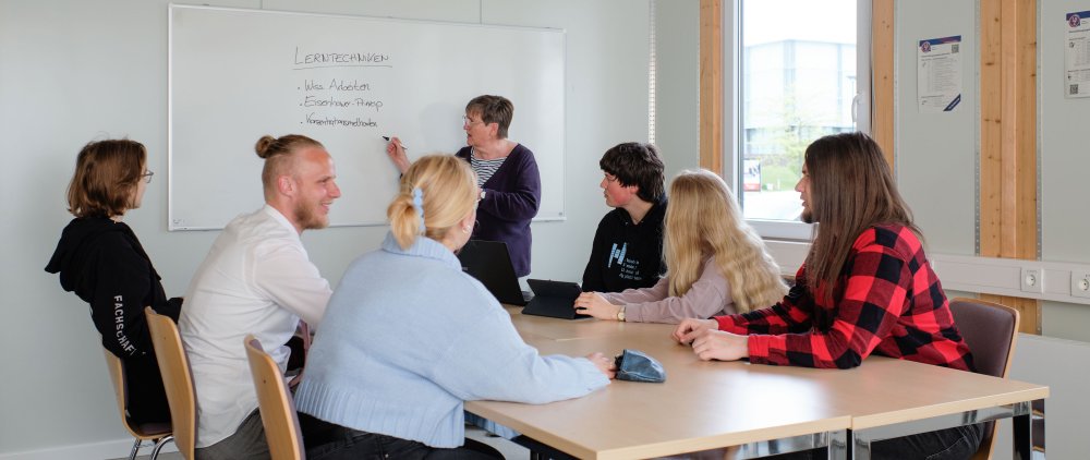 Seminarszene: Studierende sitzen um einen Tisch, vorn steht eine Dozentin am Whiteboard.