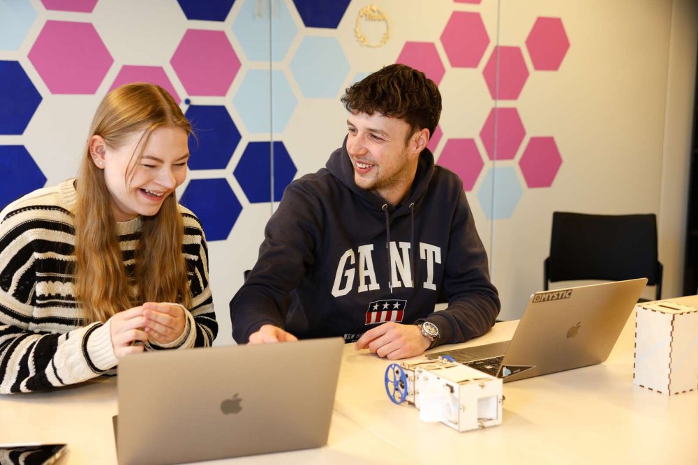 Eine Studentin und ein Student sitzen an einem Tisch, vor sich aufgeklappte Laptops, und lachen herzlich. An der Wand hinter ihnen sind rosane und blaue Kacheln.