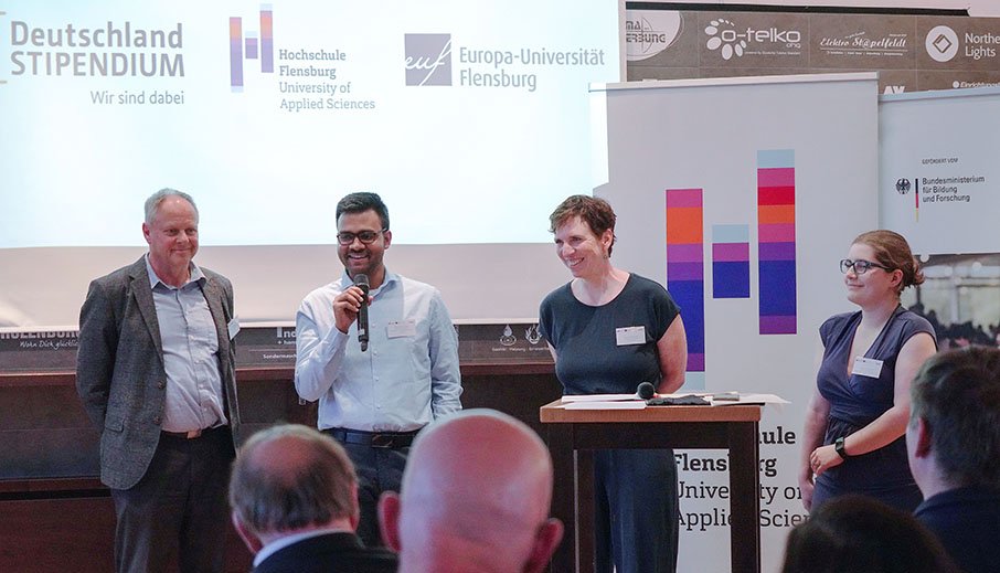 Eine Gruppe von zwei Studierenden, einem Mann im grauen Anzug und einer Frau mit kurzen Haaren steht auf einer Bühne, im Hintergrund ein Roll-Up mit dem Logo der Hochschule Flensburg, der Student spricht in ein Mikrofon.