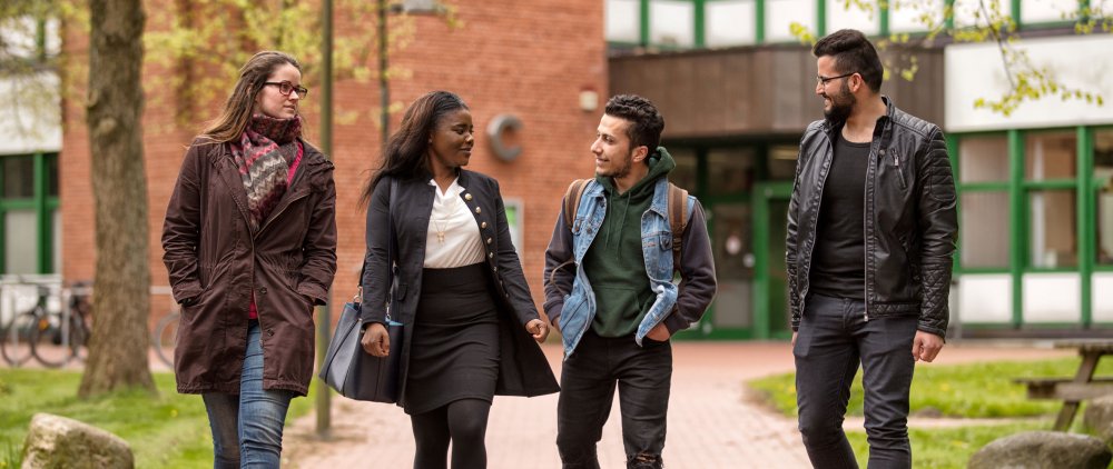 Vier Studierende, zwei Frauen und zwei Männer, laufen gemeinsam über den Campus, drei von ihnen sehen aus als hätten sie einen Migrationshintergrund.