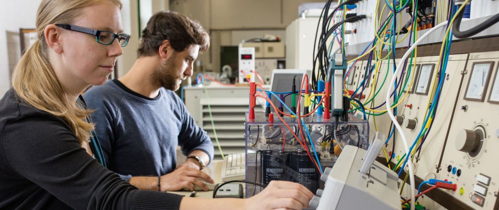 Zwei Studierende sitzen an eölektrotechnischen Geräten. Die Studentin im Vordergrund steckt Kabel um, der Student neben ihr liest in Unterlagen, die vor ihm auf dem Tisch liegen.