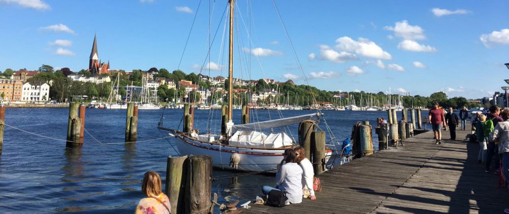 Hafen: Vorn im Bild Steg, auf dem Menschen sitzen und laufen. Hinten Wasser, Himmel und Gebäude und Bäume auf der anderen hafenseite. Auf dem Wasser ein Boot.