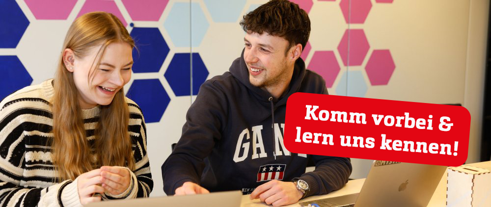 Eine Studentin und ein Student sitzen an einem Tisch, vor sich aufgeklappte Laptops, und lachen herzlich. An der Wand hinter ihnen sind rosane und blaue Kacheln. Darüber roter Störer mit Text: Komm vorbei & lern uns kennen!