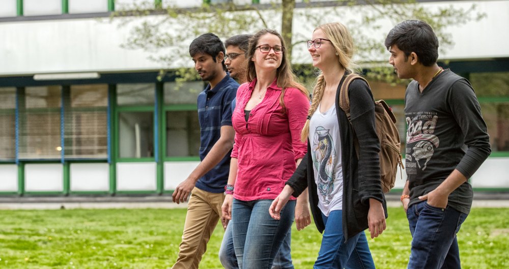 Eine Gruppe Studierender mit sichtbar unterschiedlichen Herkünften schlendert über den Campus.