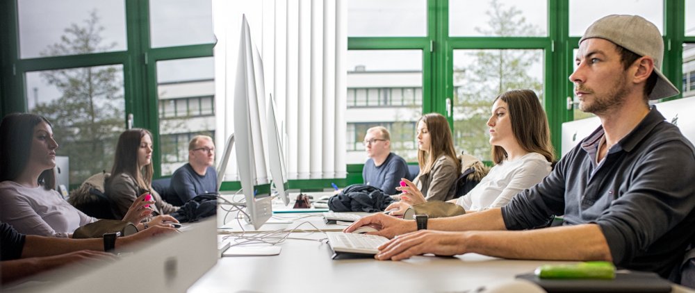 Rechts Studierende in einer Reihe an einem Tisch, sie arbeiten an Computern. Links spiegeln sie sich in den Bildschirmen.