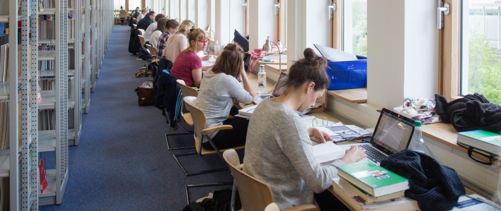 Blick auf Lernplätze in einer Bibliothek: An einer langen Tischreihe sitzen junge Menschen nebeneinander. Vor ihnen Schreibutensilien, Laptops. Links im Bild Bücherregale.