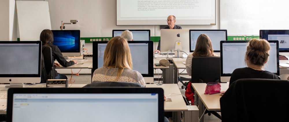 Studierende sitzen an Arbeitsplätzen mit Computern und blicken nach vorn auf den Dozenten. Das Bild ist aus einer hinteren Reihe des Raums gemacht.