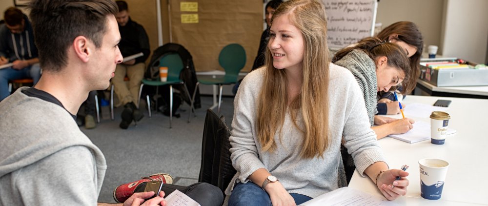 Seminarszene: Im Vordergrund ein Student und eine Studentin im Gespräch, beide haben einen Schreibblock zur Hand. Dahinter zwei Studentinnen über Unterlagen gebeugt, Stühle, weitere Menschen unscharf im Hintergrund.