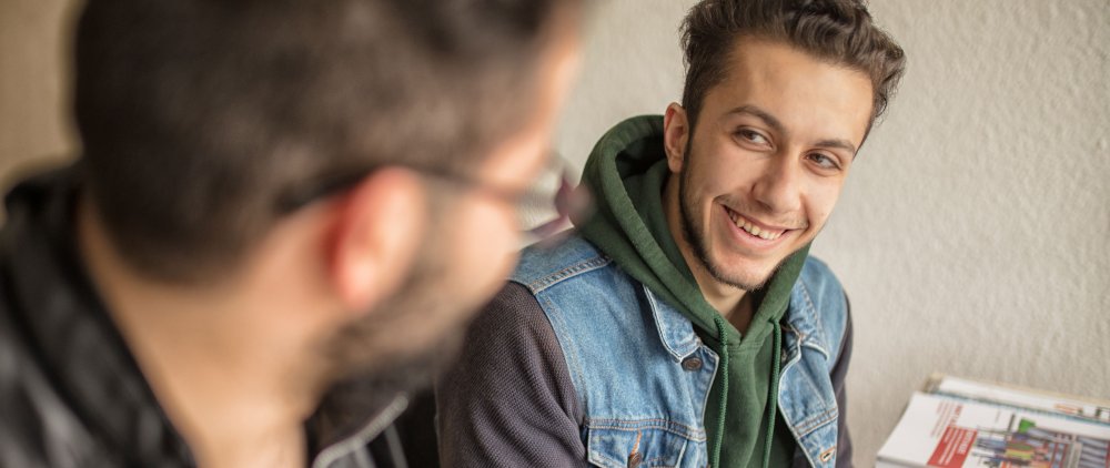 Zwei junge männliche Geflüchtete, einer ist von hinten und unscharf zu sehen, der andere schaut ihn fröhlich an und blickt dabei auch fast in die Kamera. Sie sitzen an einem Tisch, auf dem unscharf Flyer oder ähnliche bunte Dokumente zu erkennen sind.