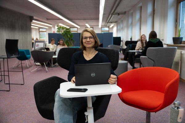Junge Frau sitzt an kleinem weißen Tisch in einer Hochschulbibliothek. Neben Ihr ist ein roter Sessel frei