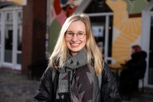 Frau mit langen blonden Haaren und einer Brille steht lächelnd vor einem Café mit bunt bemalter Ladenfront.