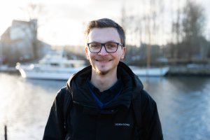 Junger Mann mit schwarzem Pulli und Brille. Im Hintergrund eine verschwommene kleine weiße Yacht in einer Hafenumgebung.