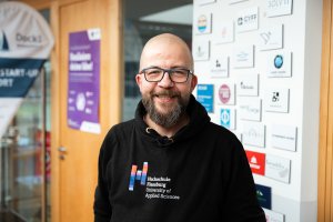 Mann mit Glatze und schwarzem Hochschule Flensburg Pullover vor Wand mit Plaketten von Firmen
