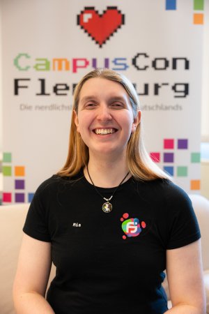 Person mit langen bloden Haaren mit buntem Fachschaft Informatik Logo auf dem T-Shirt steht vor buntem Rollup der CampusCon