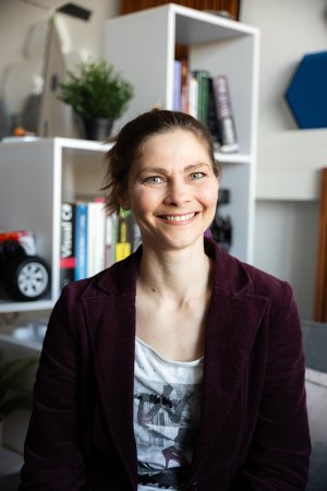 Frau mit Zopf in weinrotem Blazer. Im Hintergrund weißes Kastenregal mit vielen Büchern, Pflanzen und einem Modellauto.