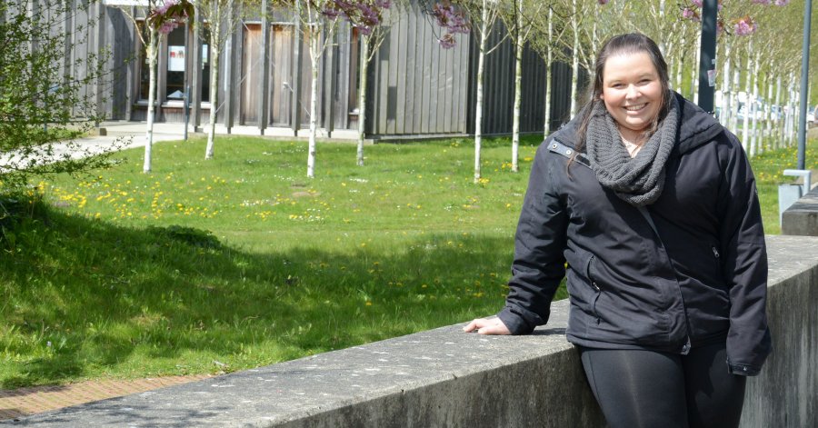 Studentin Dragana an eine niredrige Mauer gestützt auf dem Campus. Im Hintergrund sind die Campelle, Birkenstämme, eine Wiese und Kirschbaumblüten zu sehen.