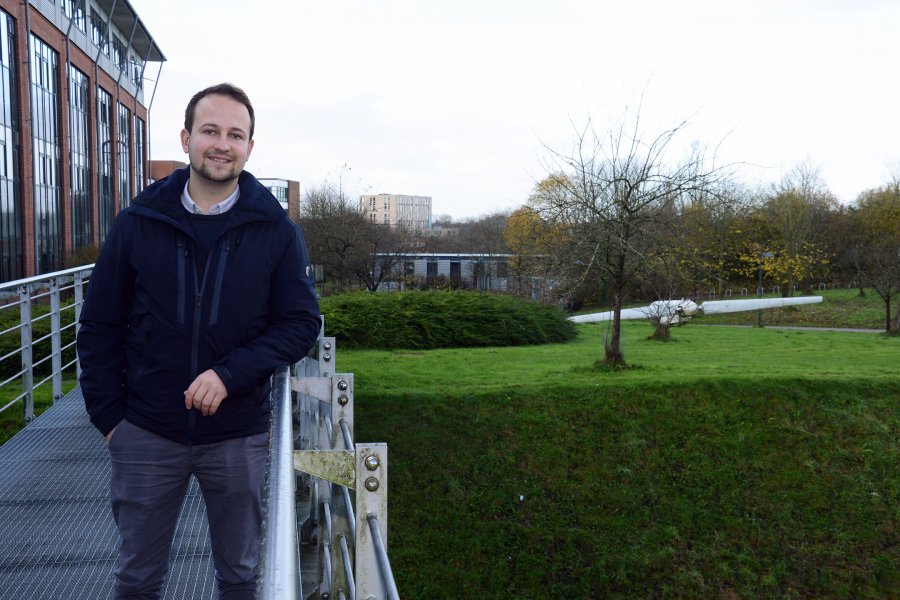 Student Jesper auf einer Brücke auf dem Campus. Hinter ihm sind ein Hochschulgebäude, eine Wiese und darauf liegend ein Propeller sichtbar.