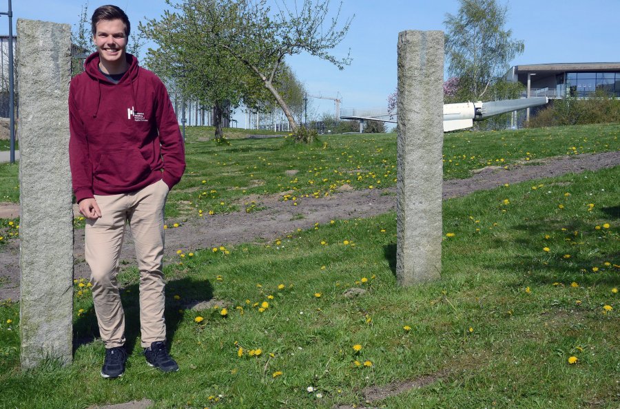 Student Patrick auf dem Campus. Im Hintergrund sind Stehlen, ein Propeller und Natur zu sehen.