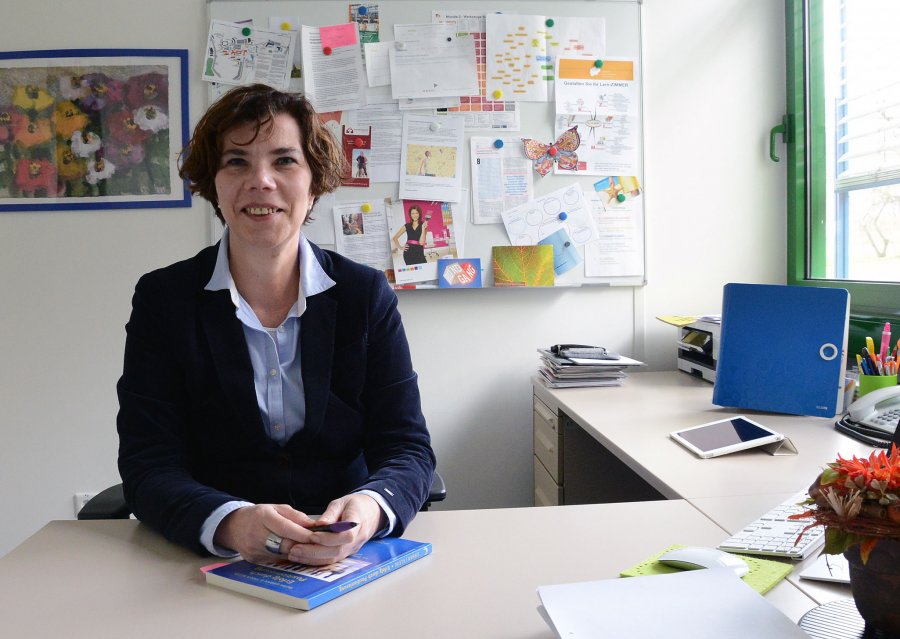 Kirsten Rohrlack an ihrem Schreibtisch. Im Hintergrund diverse Arbeitsmaterialien und ein bunt bestücktes Whiteboard.