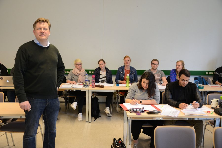 Prof. Marcus Brandenburg im Seminarraum, hinter ihm, an Tischen sitzend, Studierende.