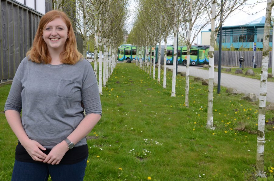 Studentin Mareike steht auf dem Campus. Im Hintergrund eine Reihe Birken und ein vorbeifahrender Bus.