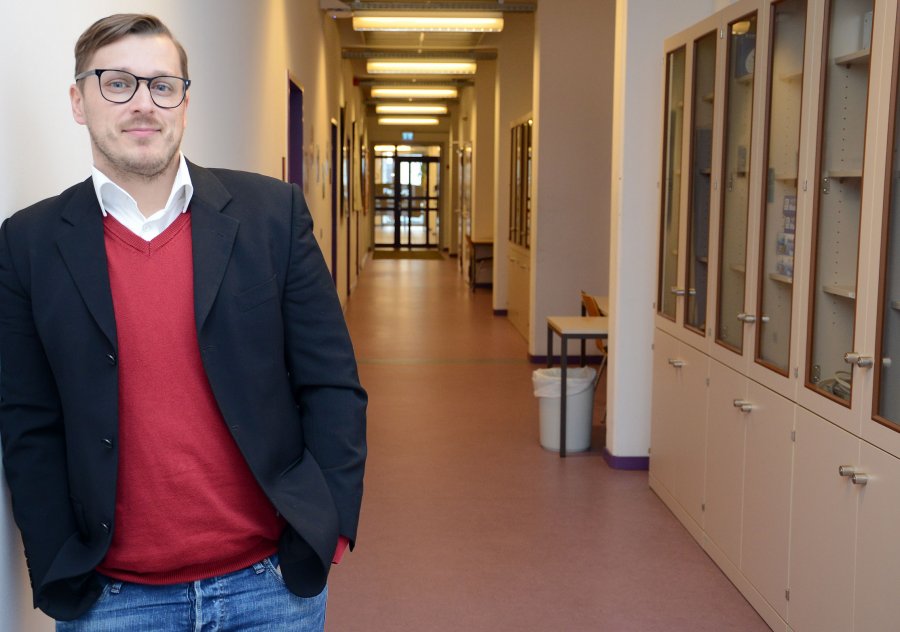 Sebastian Gajek lehnt an der Wand eines Flurs in einem Hochschulgebäude.