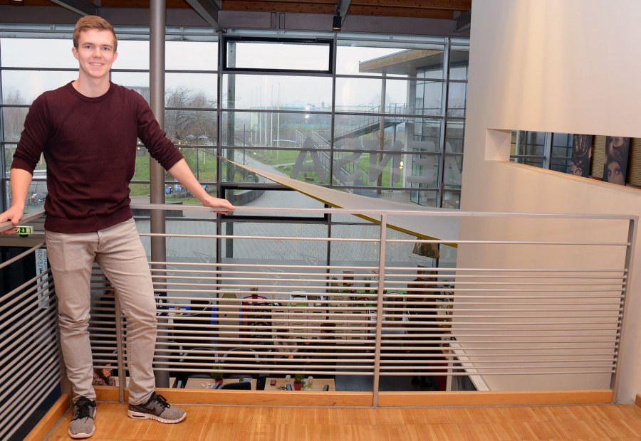 Student Tobias lehnt in einem Hochschulgebäude an einem Geländer. Hinter ihm eine Fensterwand mit Blick über den Campus.