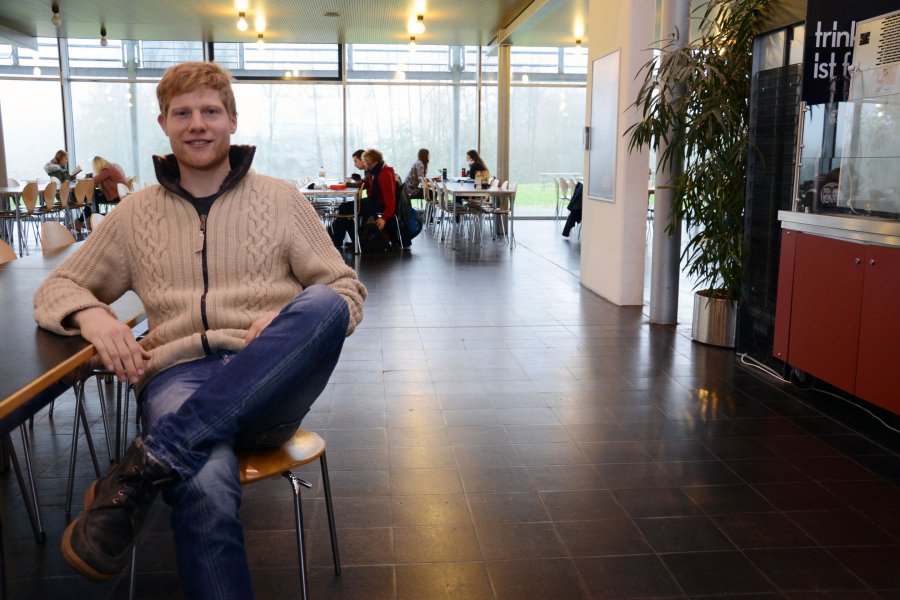 Student Jan an einem Tisch in der Mensa. Im Hintergrund ein Verkaufstresen und andere Menschen.