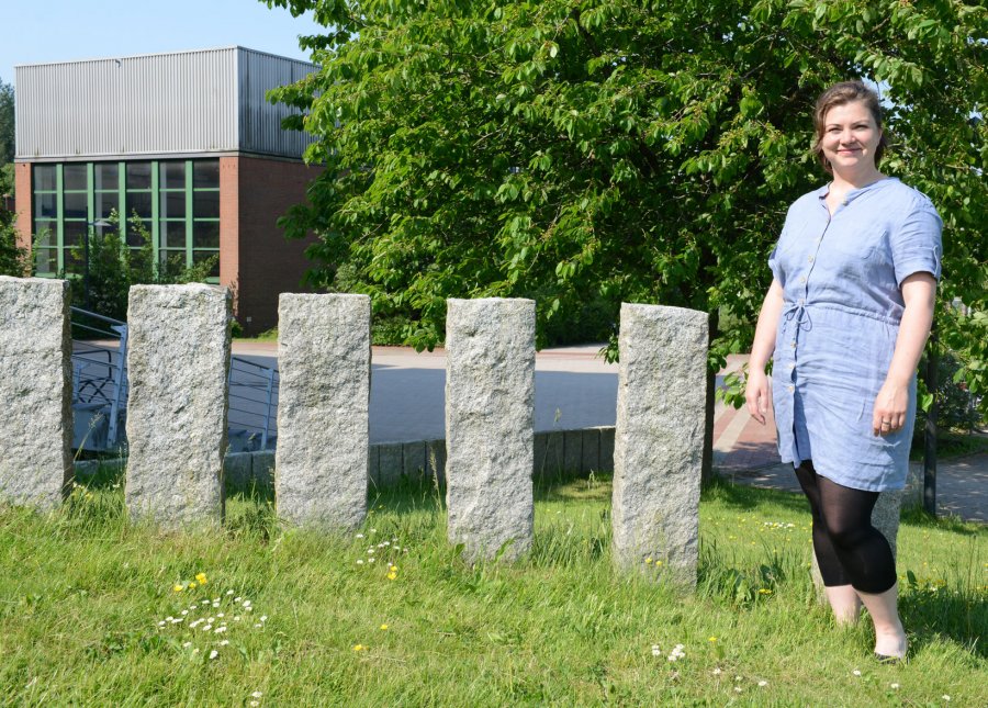 Vanessa Schomakers steht vor einer Reihe von Stehlen auf einer Wiese, im Hintergrund Bäume und ein Gebäude.