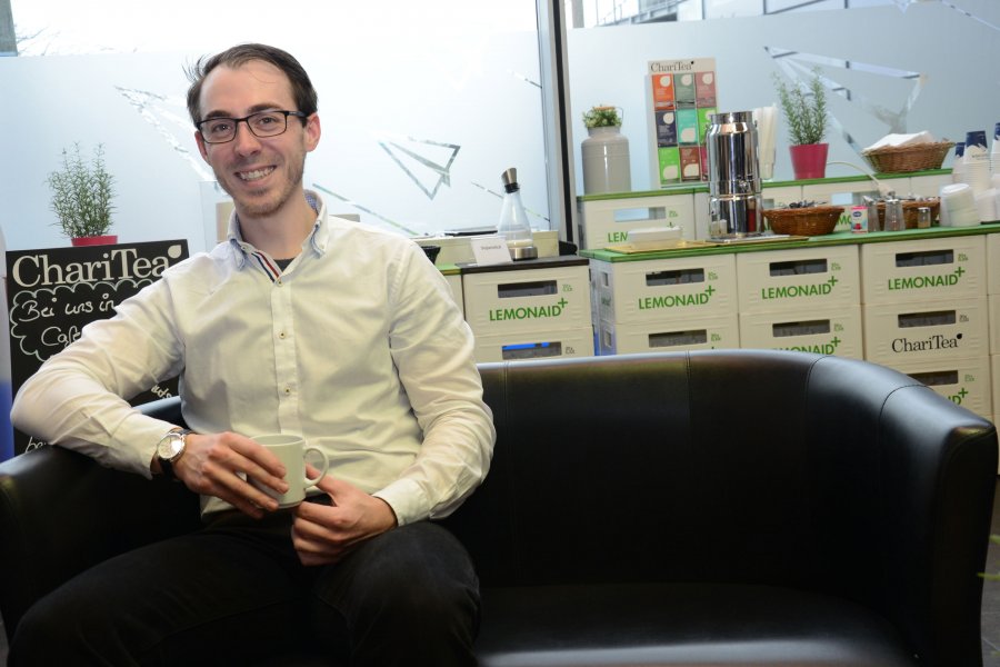 Student Martin sitzt in der Mensa auf einem schwarzen Sofa. Im Hintergrund eine aus leeren Getränkekisten gestapelte Kaffeebar.