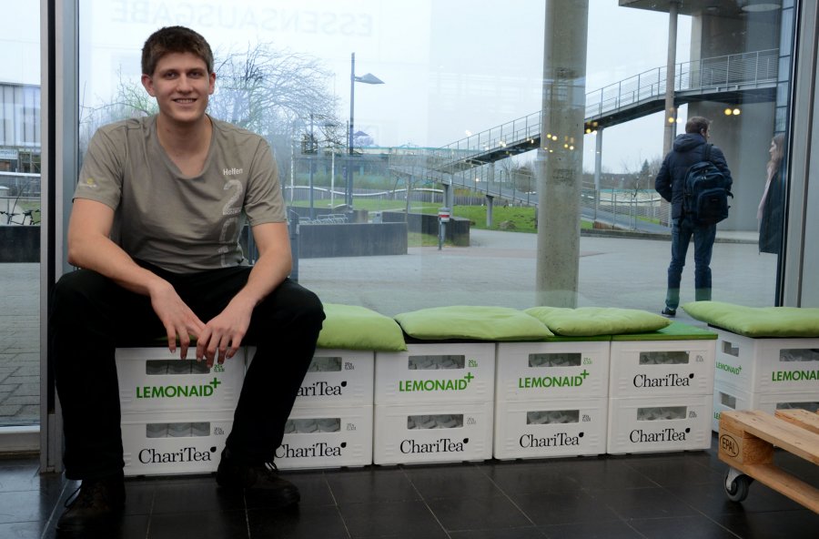 Student Benjamin sitzt auf einer Bank aus leeren Getränkekisten mit grünen Kissen darauf. Hinter ihm eine Glaswand, Blick auf einen Platz und Teile eines Gebäudes.