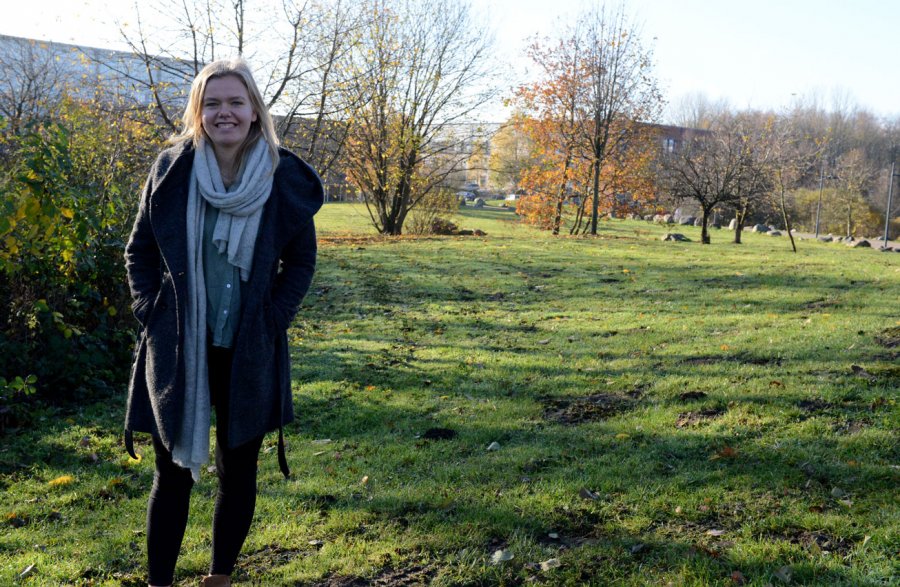 Studentin Lea steht auf einer Wiese auf dem Campus. Im Hintergrund Bäume, Büsche und ein Gebäude. Es ist sonnig, aber Herbst: Die Blätter sind schon abgefallen oder gefärbt.