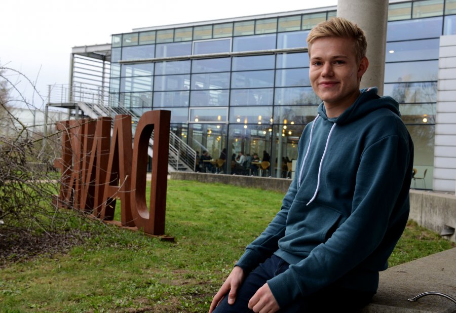 Student Tim sitzt auf einem Vorsprung, hinter ihm ist die Glasfassade der Mensa zu sehen, außerdem Rasen und eine "DANKE"-Skulptur.