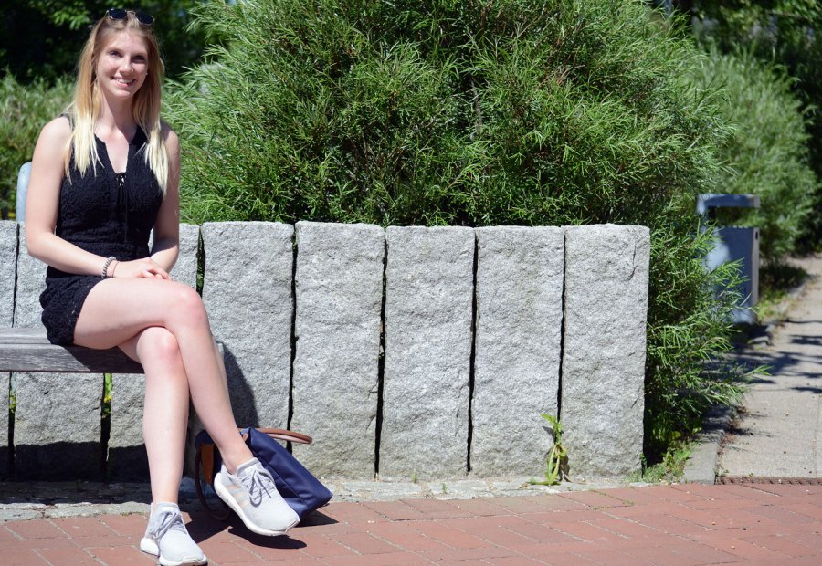 Studentin Antonia sitzt an einem Sommertag auf einer Bank. Hinter ihr eine steinerne graue Mauer, dahinter eine Hecke. Rechts im Bild der Teil eines Weges.