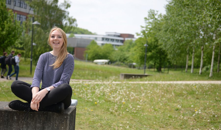 Studentin Svea sitzt auf einer Mauer auf dem Campus. Sie lacht und ihr blondes halblanges haar fliegt leicht im Wind. Hinter ihr Wiesen und Bäume. Es ist sonnig.