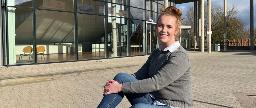 Junge Frau sitzt auf einer Mauer vor dem audimax, hinter ihr ein leerer Platz, die Glasfassade des audimax und Bäume und blauer Himmel.