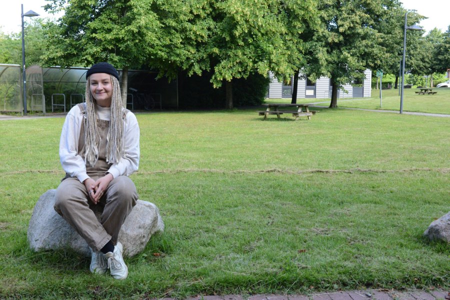 Junge Frau sitzt auf einem Stein auf einer Wiese. Sie trägt lange blonde Rasta-Zöpfe, beige Lantzhosen und ein weißen Oberteil.