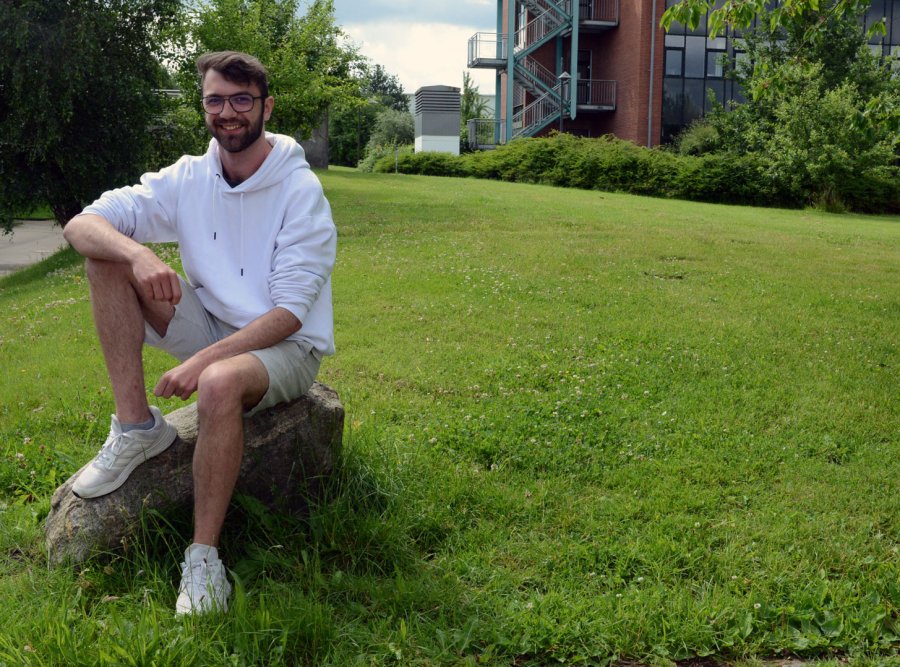 Junger Mann sitzt auf einem Stein auf einer Wiese. Hinter ihm ist ein Hochschulgebäude zu erkennen. Er trägt Shorts und ein weißes Oberteil.