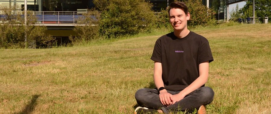 Ein junger Mann mit dunklen Haaren, schwarzem T-Shirt und schwarzer Jeans sitzt lächelnd im Schneidersitz im Gras. Im Hintergrund die Mensa auf dem Flensburger Campus.