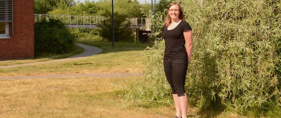 Eine junge Frau mit Brille, schwarzem T-Shirt und schwarzer Dreiviertel-Hose steht offen lächelnd auf einer Wiese vor einem Busch. Im Hintergrund die Ecke eines Backsteingebäudes der Hochschule Flensburg und eine Fußgängerbrücke aus Metall.