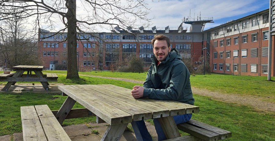 Ein Student sitzt an einer Picknickbank aus Holz und lächtelt in die Kamera. Im Hintergrund sieht man grüne Wiese, einen Baum, eine weitere Bank und ein großes L-förmiges Bachsteingebäude.