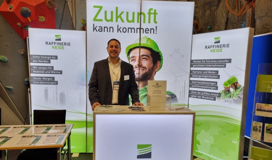 Ein Mann im Anzug steht an einem Messestand der Raffinerie Heide. Die Aufsteller sind in weiß und grün gehalten, mit dem Bild eines lächelnden Mannes mit grünem Bauhelm und der Überschrift "Die Zukunft kann kommen!".