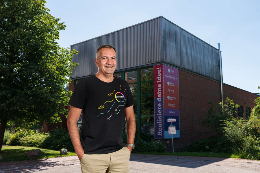 Ein Mann im mittleren Alter im schwarten T-Shirt mit buntem Aufdruck "Covestro". Im Hintergrund ein Backsteingebäude mit Glasfront und einem buten Banner mit Aufdruck "Realisiere deine Idee!".