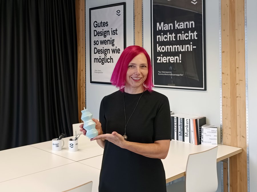 Eine Frau mit leuchtend pinken Haaren und schwarzem Kleid steht vor einem Schreibtisch, an der Wand hinter ihr zwei Poster mit Text: "Gutes Design ist so wenig Design wie möglich" und "Man kann nicht nicht kommunizieren!".