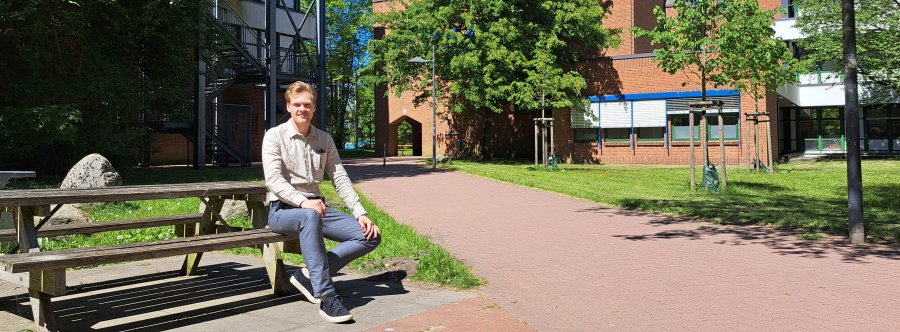 Ein junger Mann sitzt im strahlenden Sonnenschein auf einer Picknickbank auf dem Flensburger Campus, im Hintergrund Bäume, Backsteingebäude und blauer Himmel.
