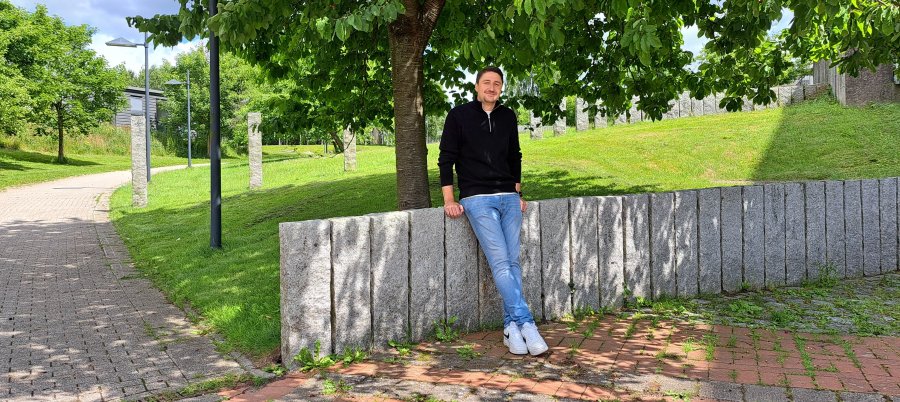 Ein junger Mann im schwarzen Troyer, Jeans und weißen Sneakern sitzt auf einer niedrigen Steinmauer und lächelt in die Kamera. Im Hintergrund eine grüne Wiese, Bäume und Steinstelen.